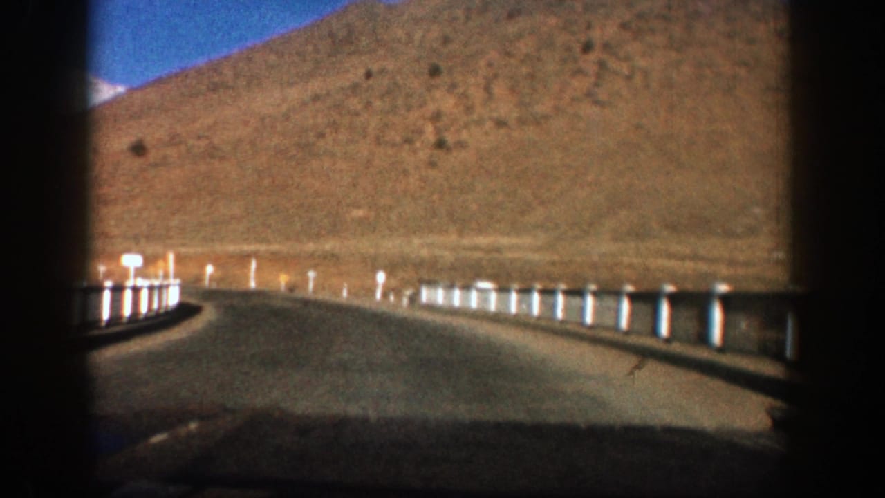 1961 Aspen, Colorado road view from a moving car, showing a curve, roadside posts, dry slopes, and blue sky.