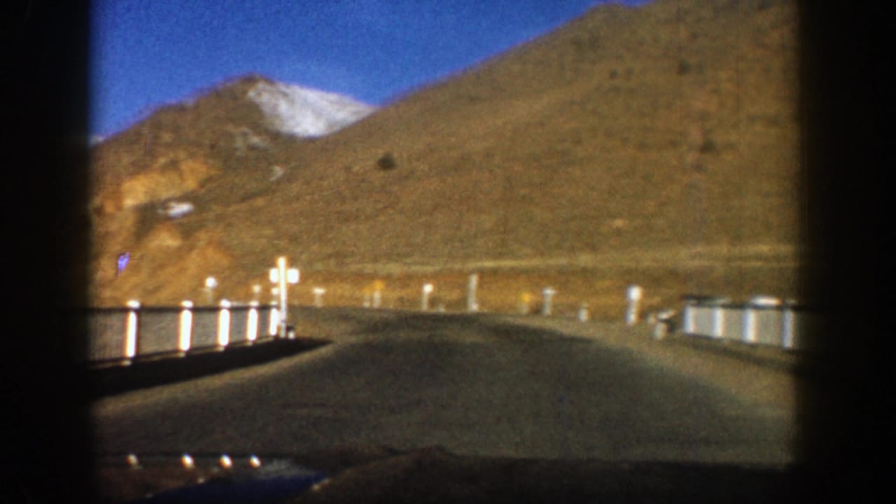 Mountain road curving past guardrails with a snow-tipped peak in the distance.