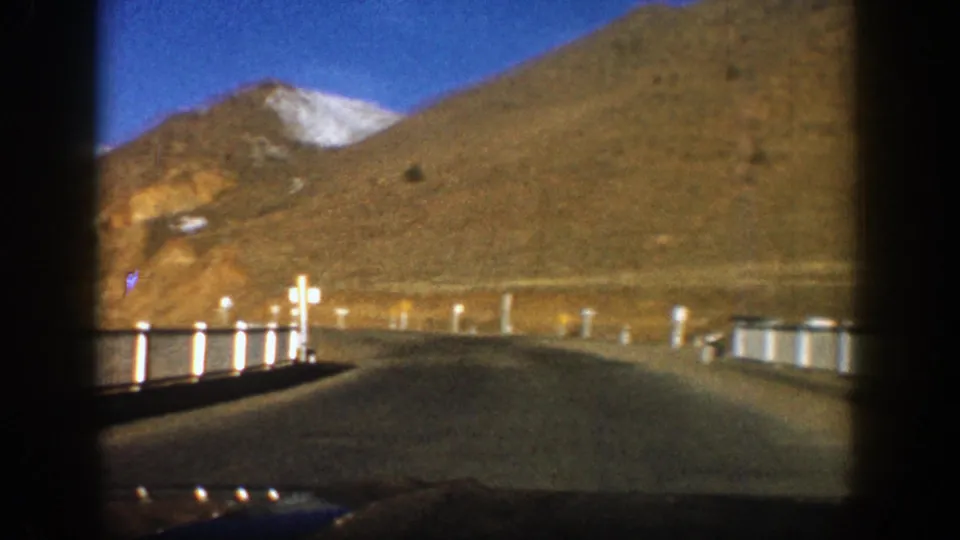 Moving-car view in Aspen, Colorado, with a winding road, guardrail posts, dry mountain slopes, and a snow-tipped peak.