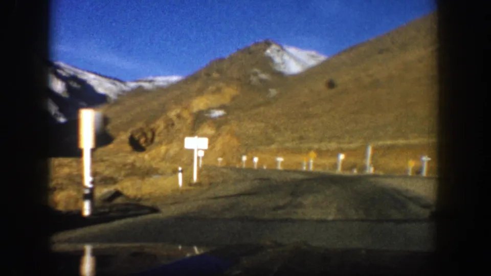 1961 Aspen mountain-road frame with roadside signs, white posts, arid slopes, and snow-capped peaks beyond.