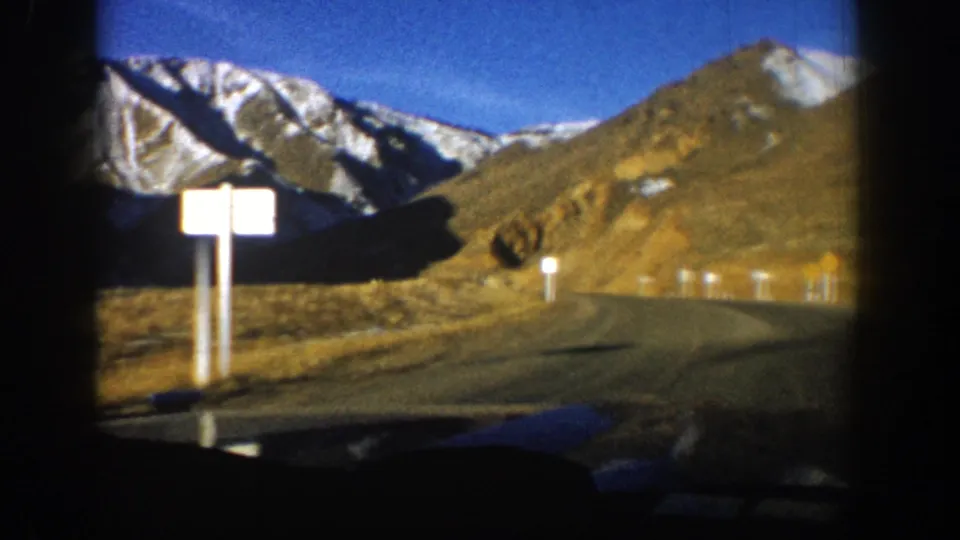 Aspen, Colorado home-movie road view showing a curve, road signs, white posts, dry slopes, and snow-streaked peaks.