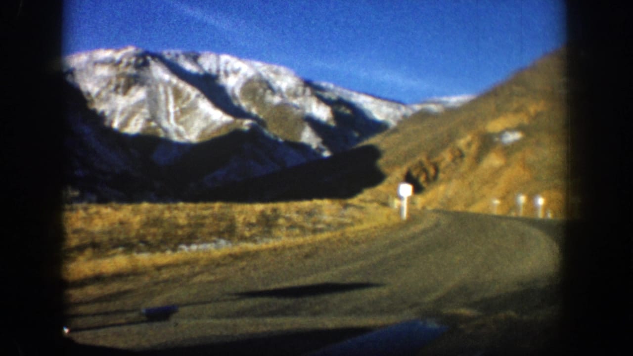 1961 Aspen-area 8mm view from a moving car on a paved road toward snow-dusted Colorado mountains.