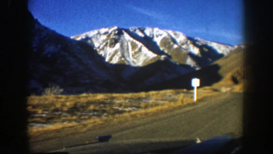 Passenger-view 8mm frame of a Colorado mountain road with dry hillsides and snow-striped peaks near Aspen in 1961.