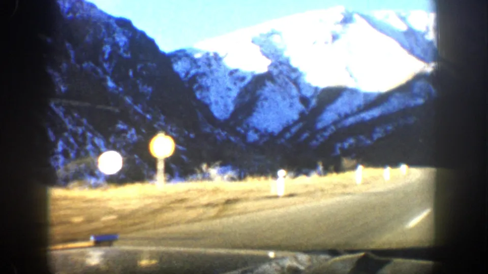 Overexposed 8mm frame of a road through snowy Colorado mountains, with roadside markers and dashboard edge visible.