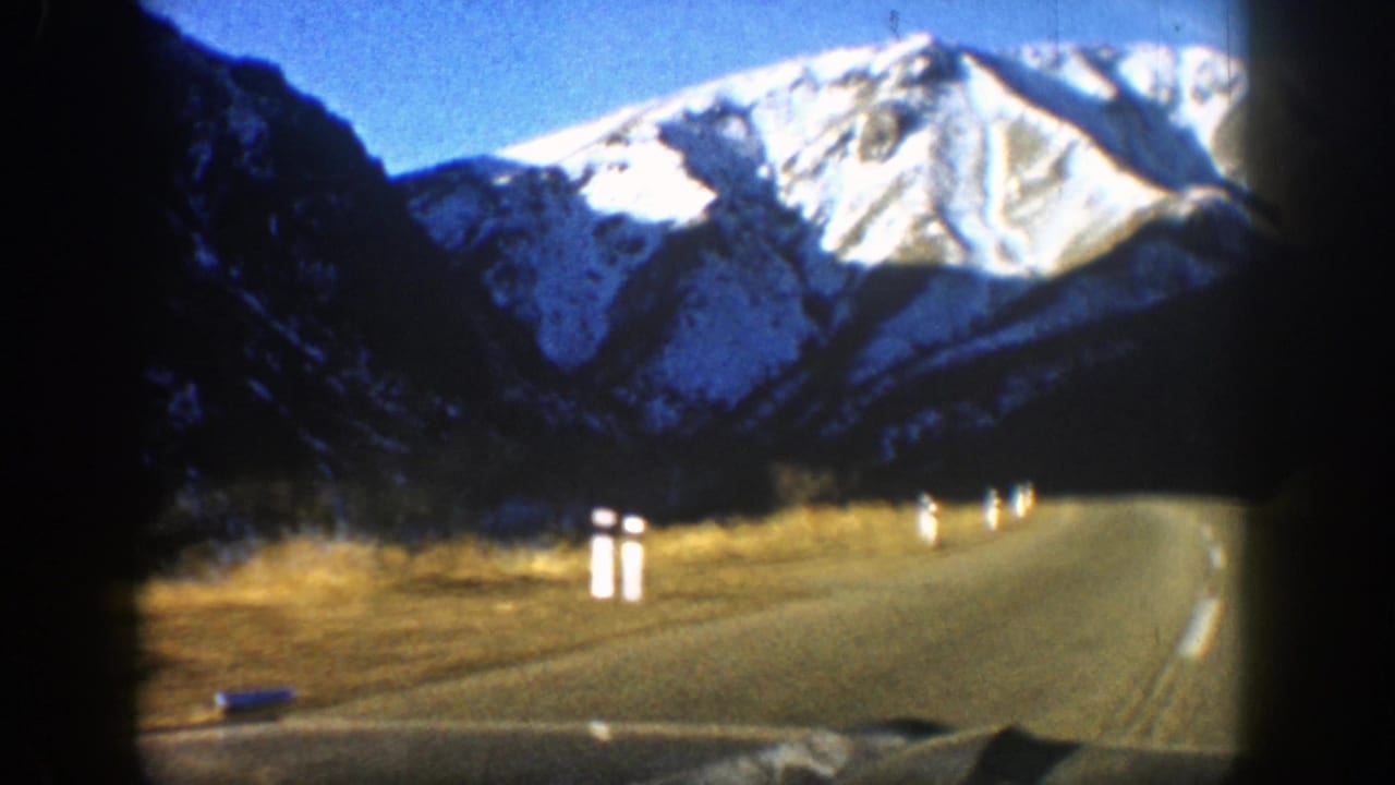 1961 Aspen home-movie frame showing a mountain road curving toward snow-dusted peaks under a blue sky.