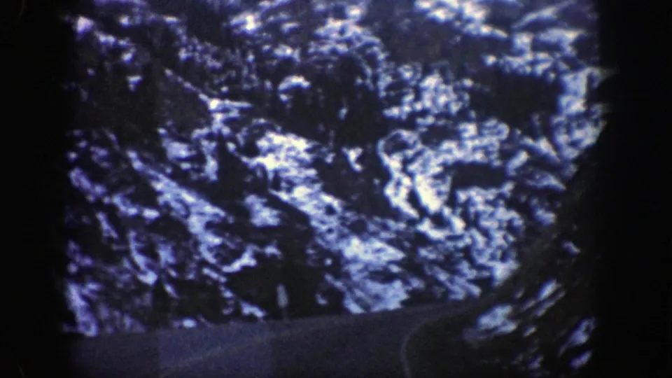 1961 8mm view of a winding mountain road near Aspen, Colorado, bordered by dark canyon slopes and patches of snow.