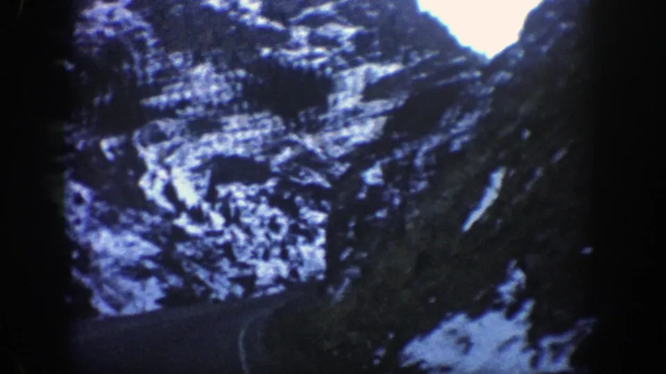 A vehicle-view shot along a narrow mountain route in the Aspen area, with snow on the ridges and a bright opening ahead.