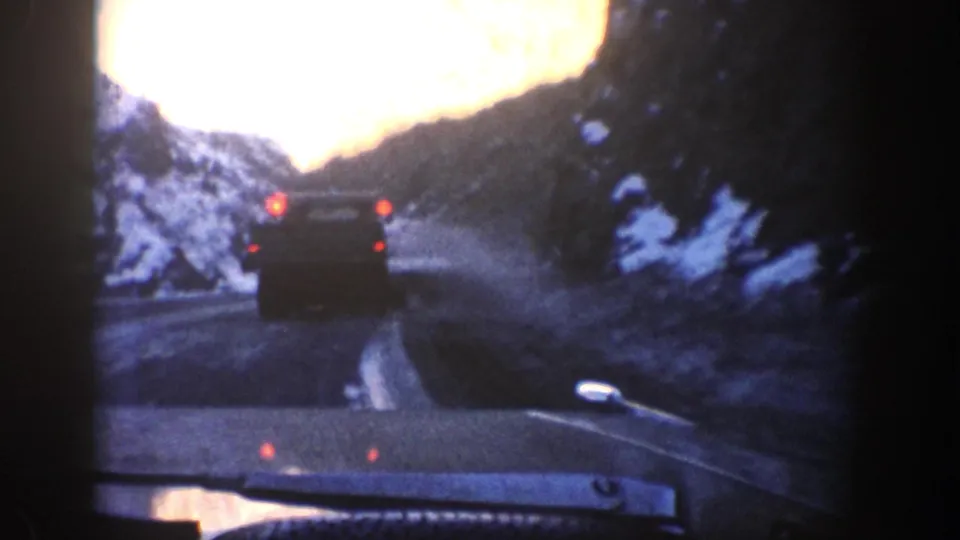 From inside a vehicle, a dark car drives ahead on a snow-lined Aspen-area mountain road, its red taillights visible in soft 8mm color.