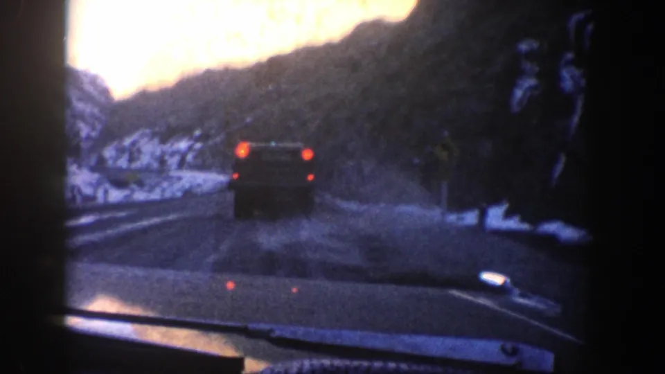 1961 8mm frame showing a car ahead on a wet mountain road with snowbanks and dark slopes near Aspen.