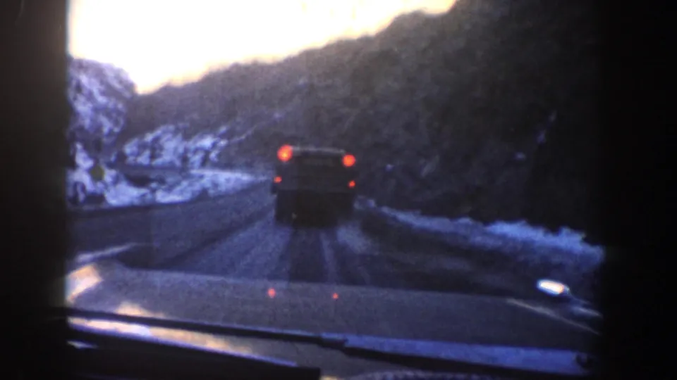 End frame of a 1961 Aspen-area road view, with a vehicle ahead and snowy mountain roadside visible through the windshield.
