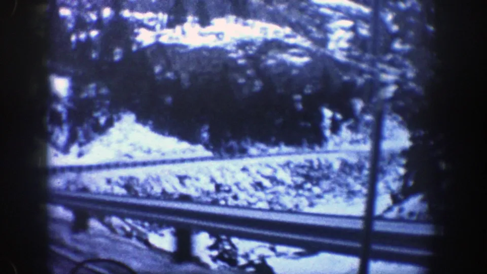 1961 Aspen 8mm frame with a roadside guardrail, snowy embankment, trees, and a rock slope seen from a moving vehicle.