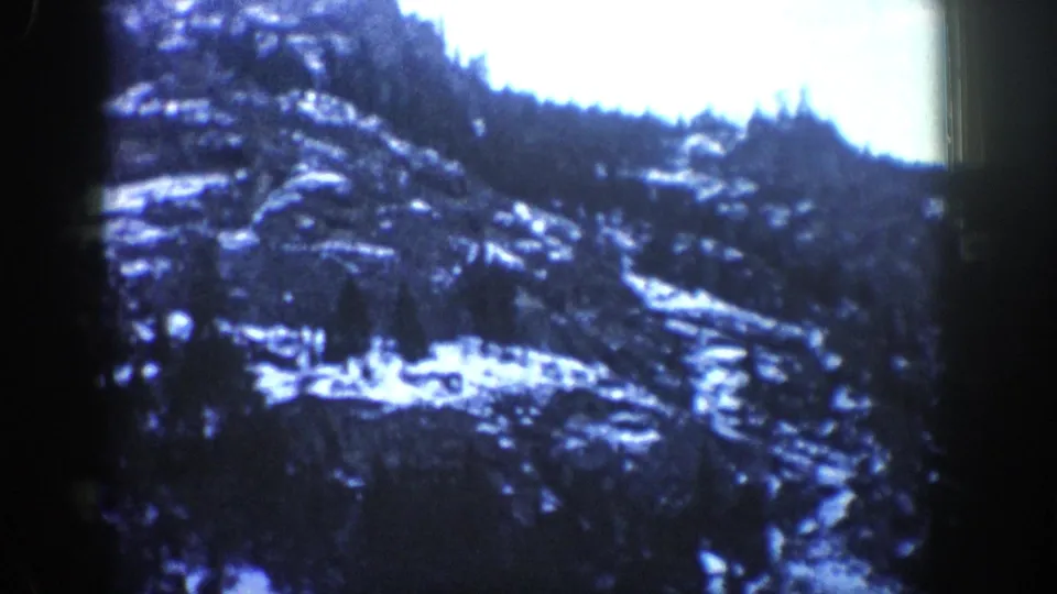 1961 Aspen, Colorado home-movie frame showing a snowy rocky slope and evergreen treeline from a moving car.