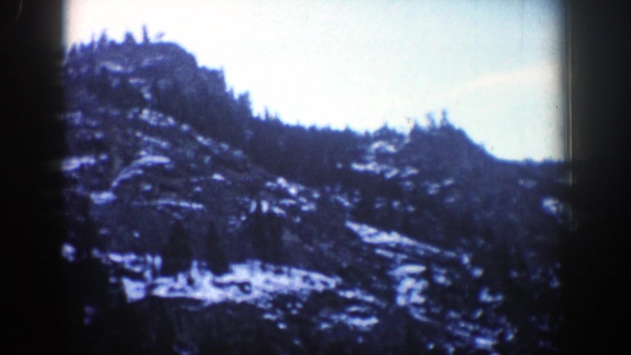 Rocky mountain ridge with snow patches and dark trees beneath a pale sky.