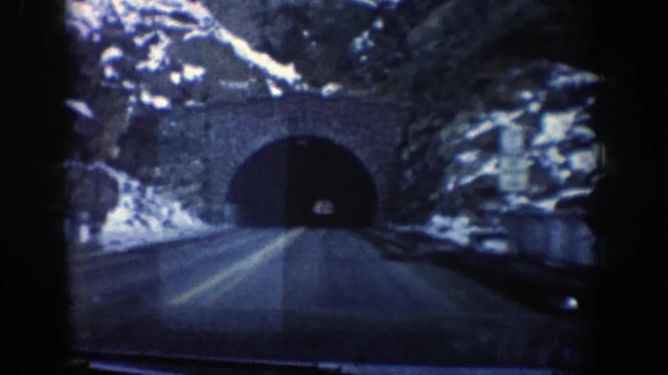 1961 Aspen-area mountain drive frame showing a two-lane road leading toward a stone-arch tunnel under snowy rock slopes.