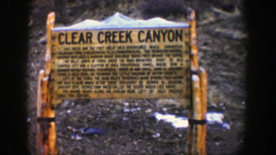 Close 8mm frame of the Clear Creek Canyon roadside marker, with large title lettering, wooden supports, and snow on the ground.