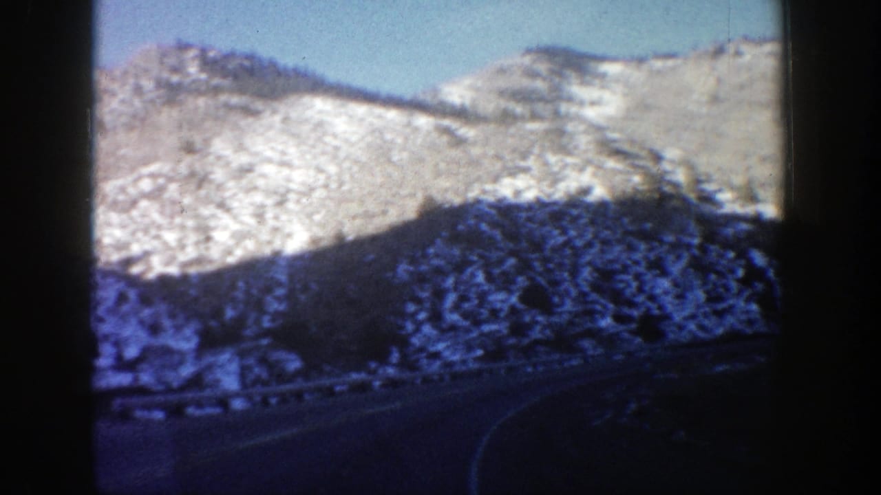1961 Aspen, Colorado home-movie frame of a curved mountain road below snowy rocky slopes, viewed from inside a car.