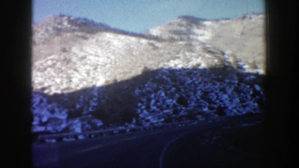 1961 Aspen, Colorado home-movie frame of a curved mountain road below snowy rocky slopes, viewed from inside a car.