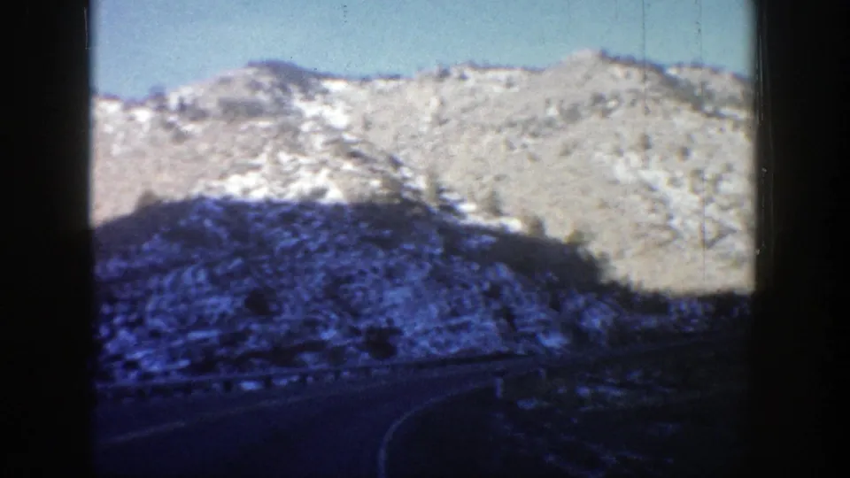 Passenger-view 8mm frame from Aspen, Colorado, showing a bend in the road beneath pale snowy hills and a clear sky.