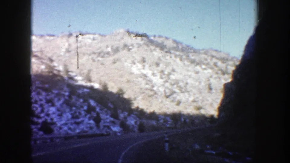 A 1961 home-movie view in Aspen, Colorado, with a snowy rocky hillside, blue sky, and a dark road curve below.