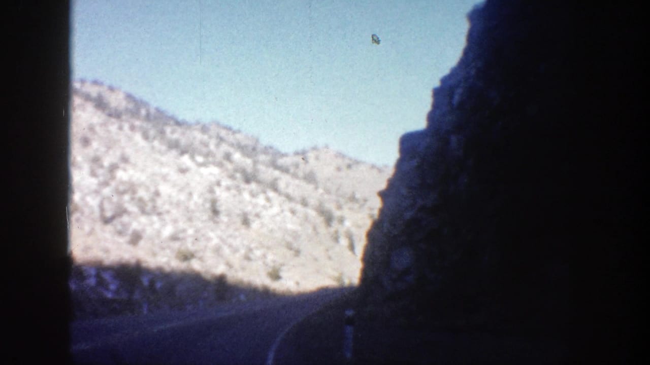Sunlit tan mountains and blue sky seen from a dark mountain road.