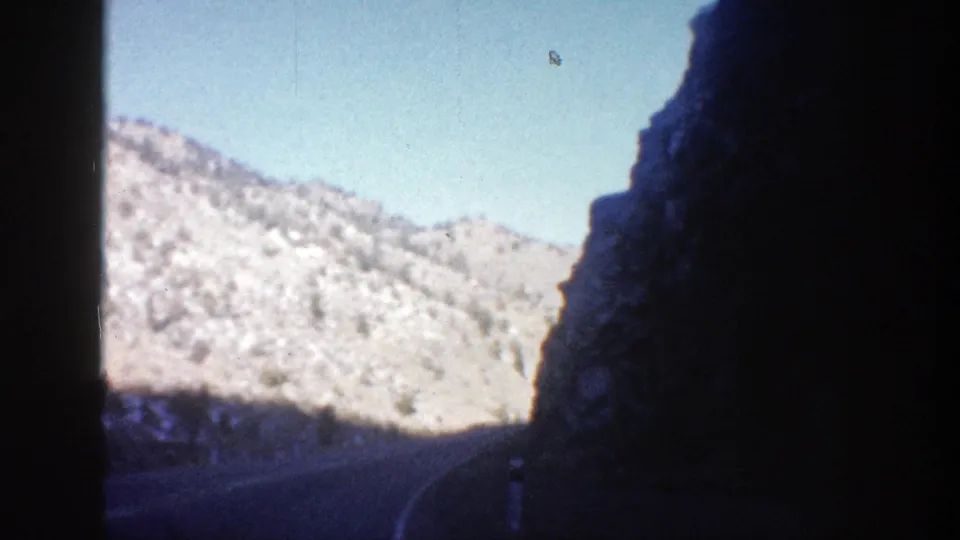 1961 Aspen-area mountain road view with tan rocky slopes, blue sky, and deep shadow along the roadway.