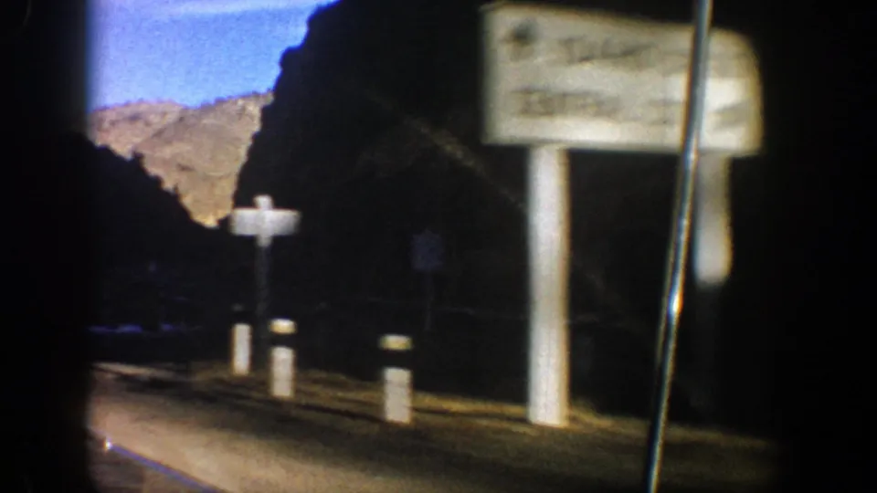 Roadside sign, white posts, and dark canyon wall appear during the 1961 Aspen-area mountain drive.