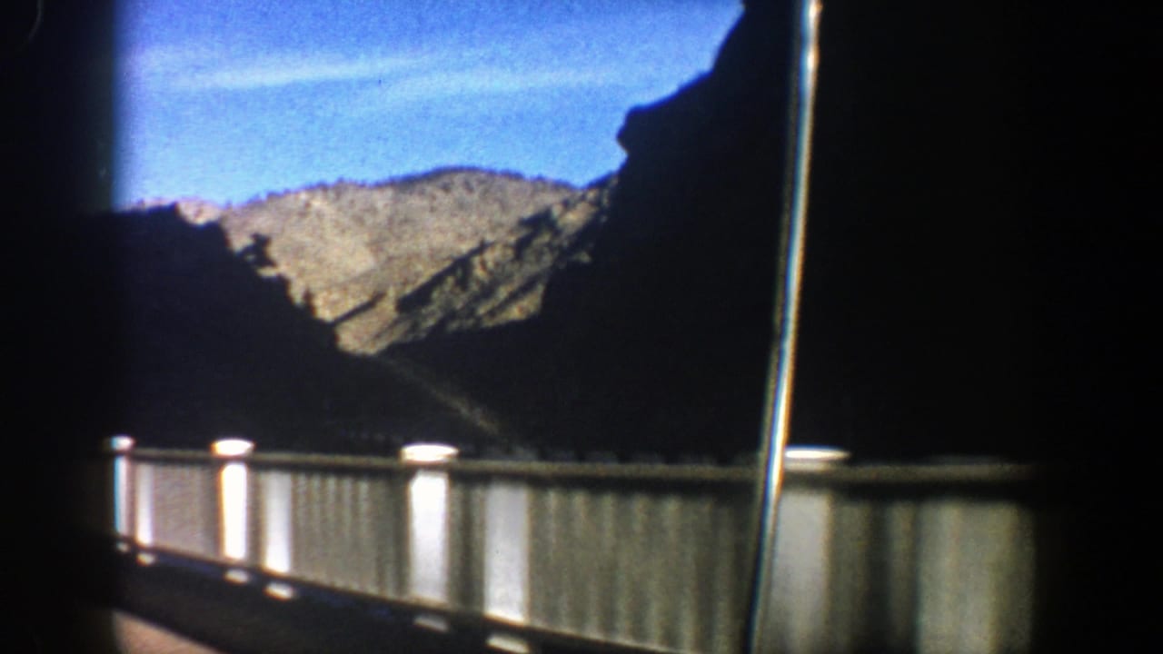White bridge railing seen through a vehicle window with rocky mountains and blue sky beyond.