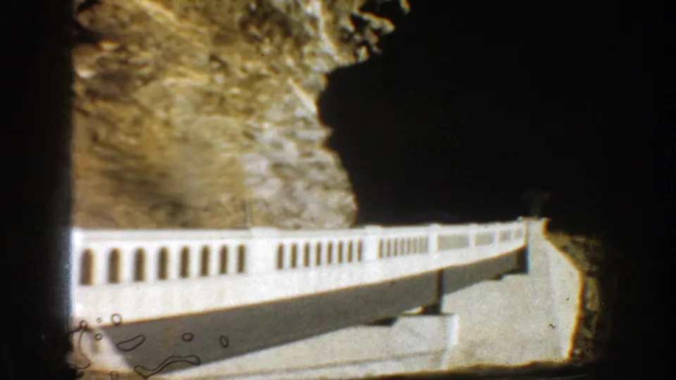 A close 8mm view of the white bridge railing and adjacent cliff, with dark gate edges and small film marks visible.