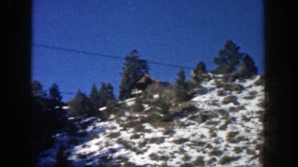 1961 Aspen-area frame showing evergreen trees and a small hillside structure above patchy snow under a deep blue sky.