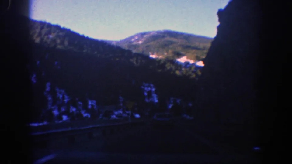 1961 Aspen-area landscape frame with dark foreground trees, snow patches, and distant mountain ridges seen from the road.