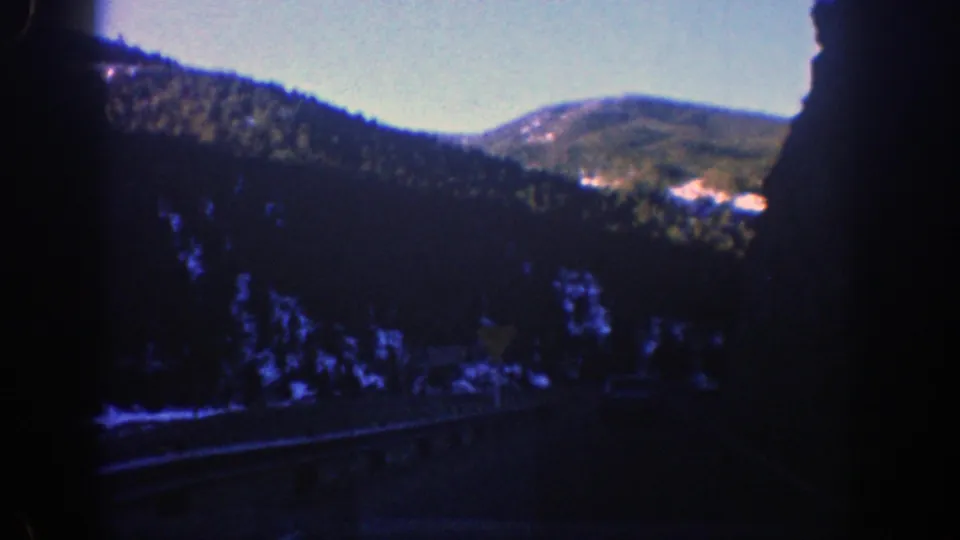 1961 Aspen, Colorado home-movie frame showing evergreen hills and pale mountain patches from a moving car window.