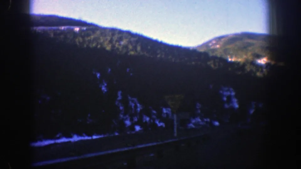 Moving-car view in Aspen, Colorado, with shadowed roadside trees, mountain hills, and a briefly visible yield sign.
