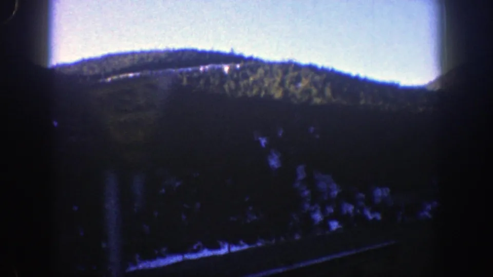 1961 8mm mountain-road frame showing a forested Aspen hillside through heavy window shadow and blue-shifted film color.