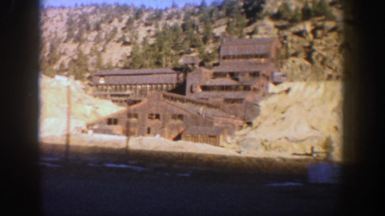 1961 Aspen home-movie frame showing weathered mine structures below a rocky, tree-covered hillside with snow on the ground.