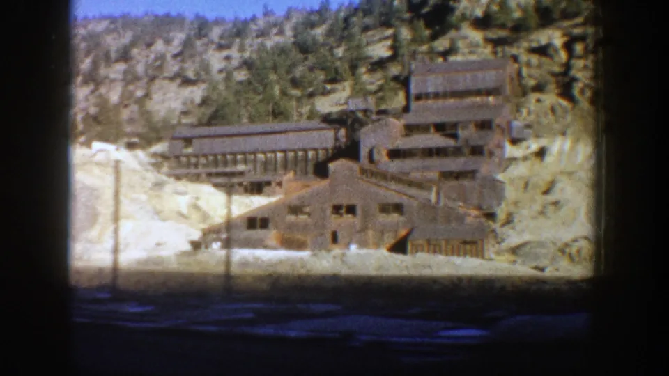 1961 Aspen frame of an abandoned-looking mining complex with long timber buildings, snow patches, and trees on a rocky slope.
