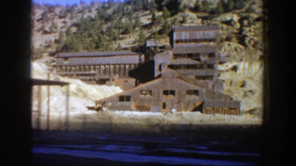 1961 Aspen view of weathered mine buildings and pale snowy or tailings-covered ground beneath a wooded rock face.