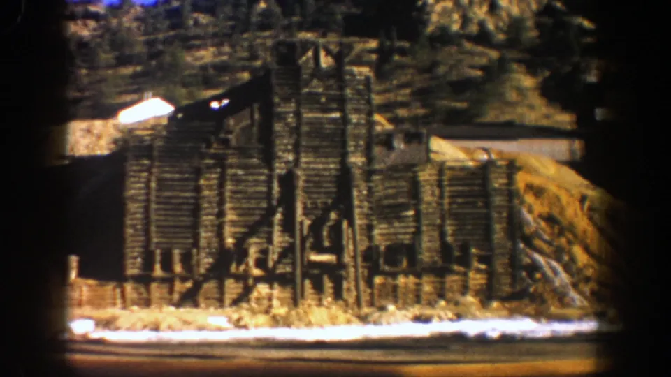 1961 Aspen frame showing a darker timber industrial structure near snow patches and a rocky hillside, with the camera passing by.