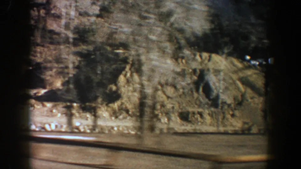 Aspen-area home-movie frame showing a shadowed roadside, tan mountain base, and bare trees with visible 8mm frame edges.