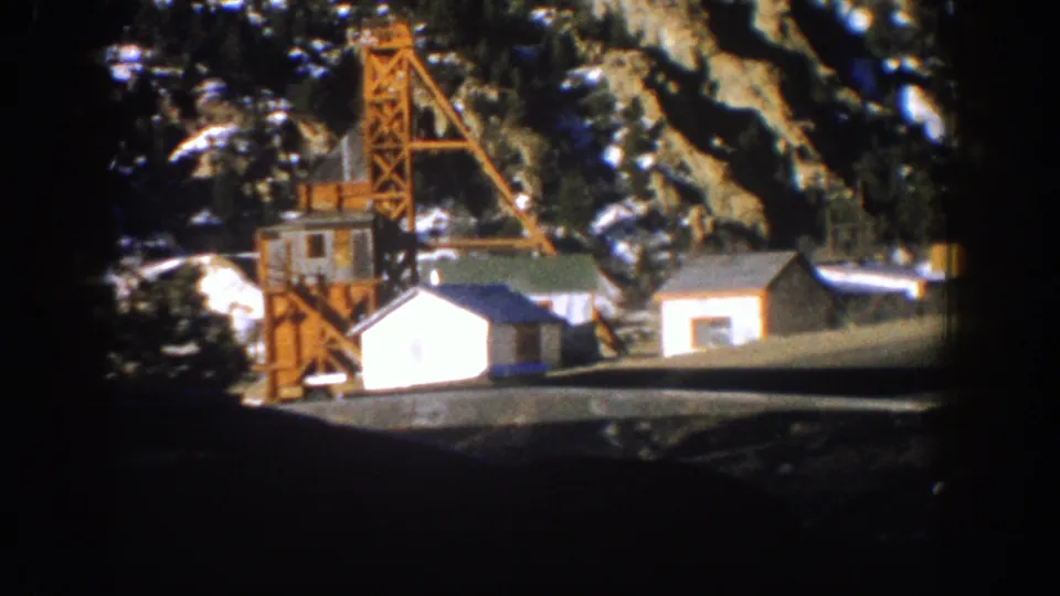 1961 Aspen, Colorado frame with small white utility buildings, an orange crane-like structure, evergreens, and rocky mountainside.