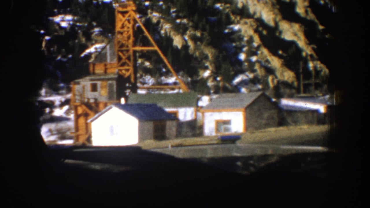 1961 Aspen, Colorado home-movie frame showing small wooden buildings and a derrick-like tower below snow-dusted evergreens.