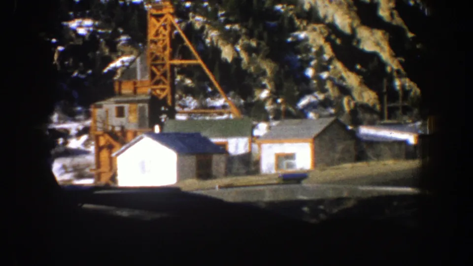 1961 Aspen, Colorado home-movie frame showing small wooden buildings and a derrick-like tower below snow-dusted evergreens.