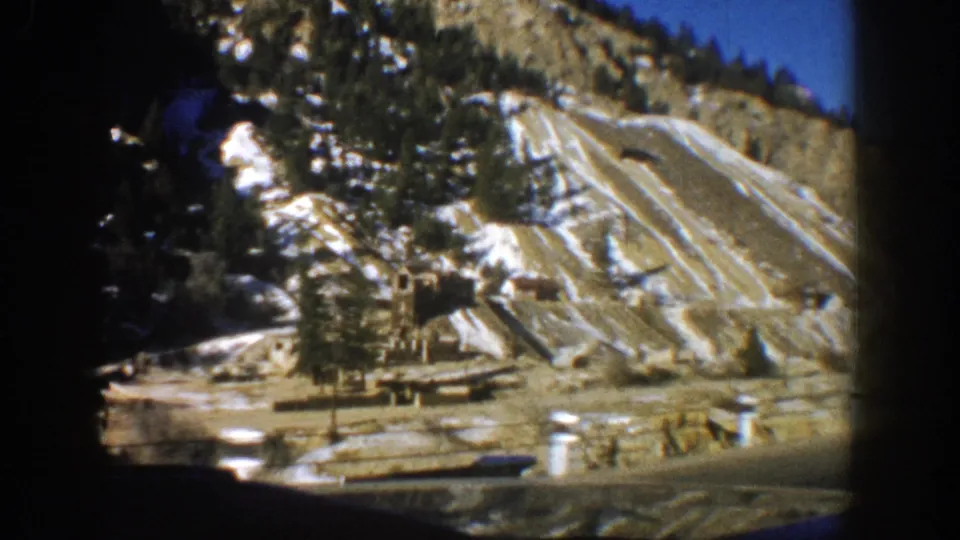 Car-framed 1961 Aspen view of exposed terraced earth, snow patches, trees, and small roadside structures on a mountain slope.