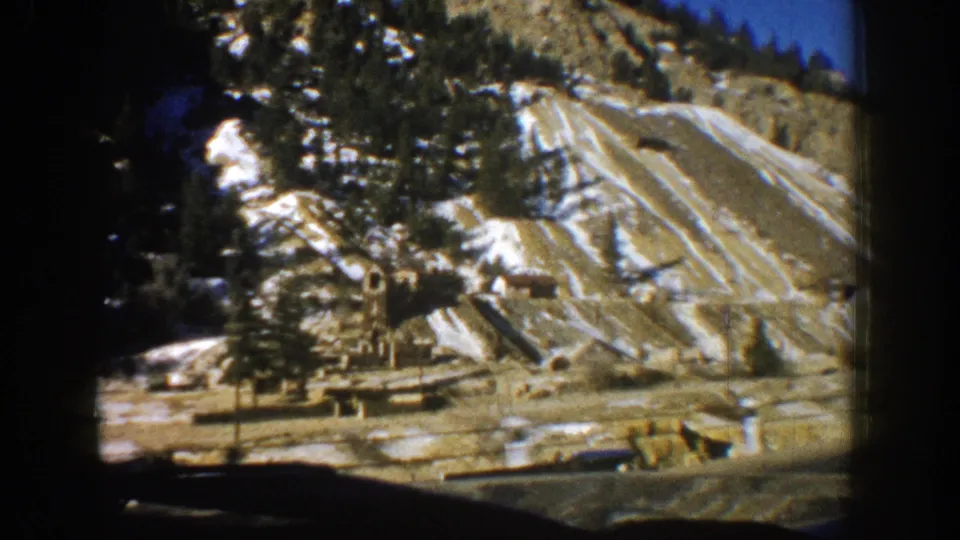 1961 Aspen car-window view of a steep terraced slope with exposed earth, snow streaks, trees, and mountain-road foreground.