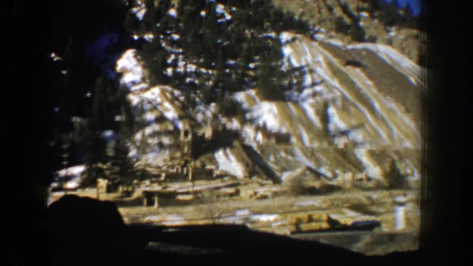 Final 1961 Aspen frame showing a dark car interior edge against a rocky, snow-patched mountain slope with evergreens.