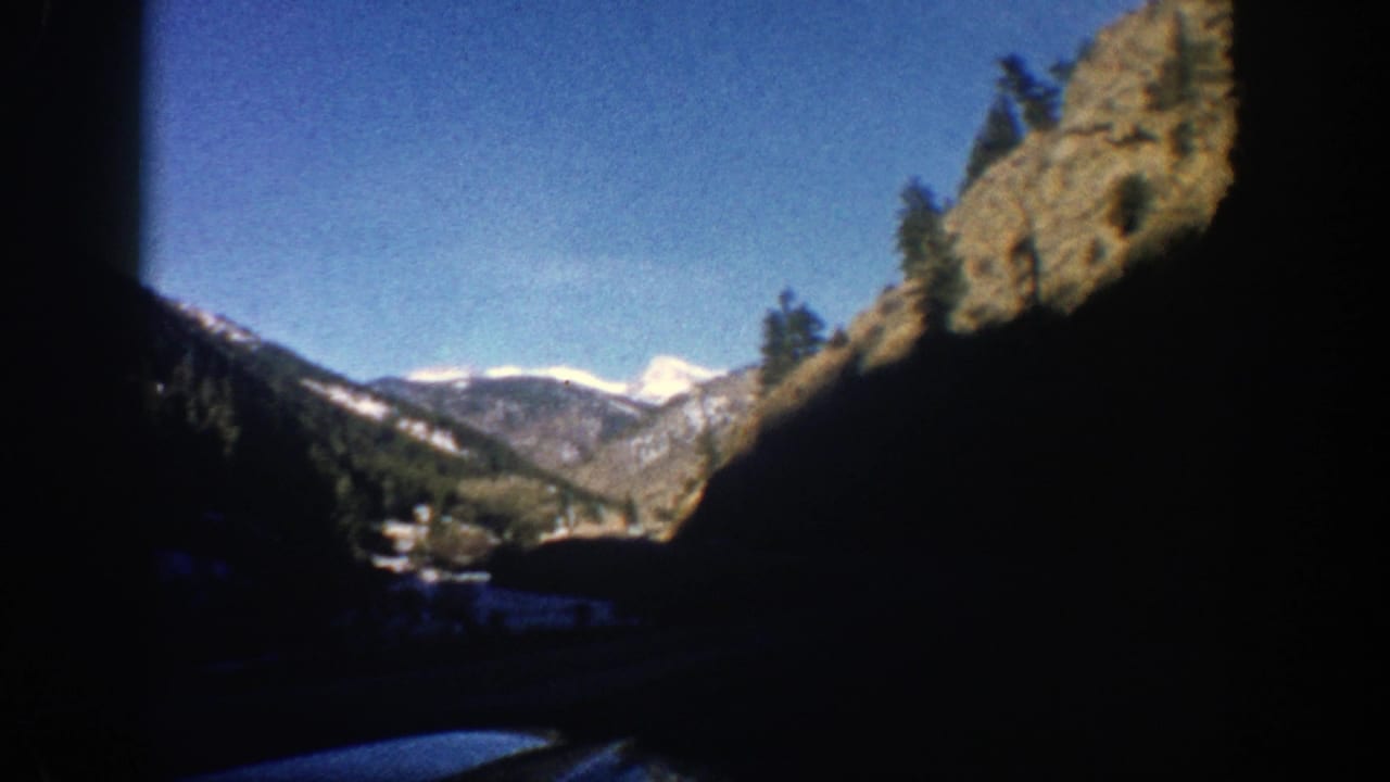 Mountain valley seen from a moving car with dark window edges and snow-capped peaks in the distance.