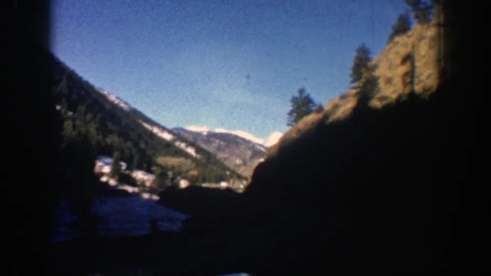1961 Colorado mountain roadside view from a moving vehicle, with dark foreground framing, trees, snow patches, and distant ridges.