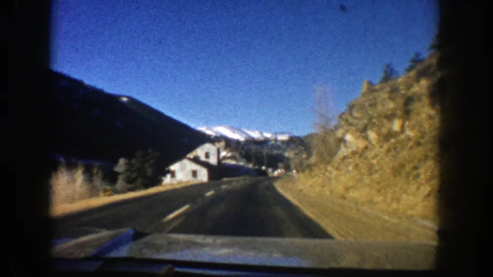 Aspen-area 1961 road view from a car, showing pavement, rocky roadside, a small house, evergreens, and snow-capped mountains.