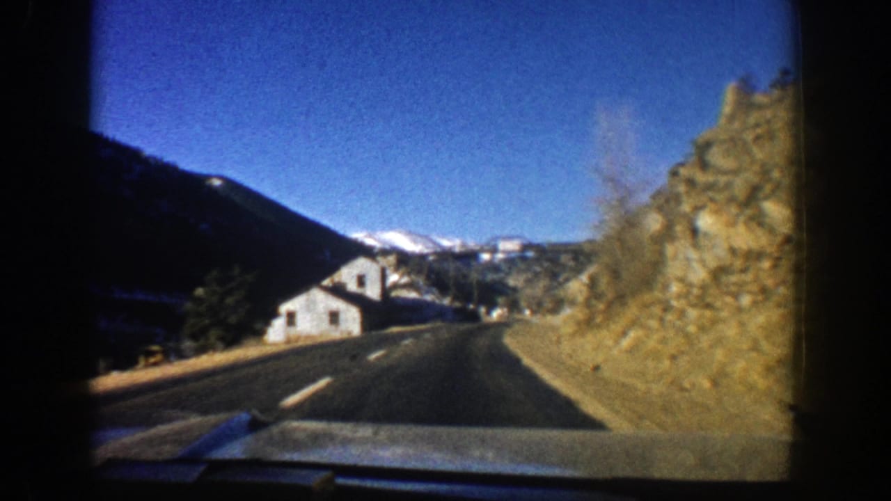 Road view toward a white building between rocky slopes and distant snowy mountains.