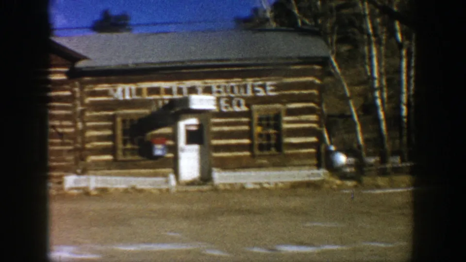 1961 Aspen-area pass-by of a rustic log cabin exterior with a partially readable painted sign and roadside foreground.