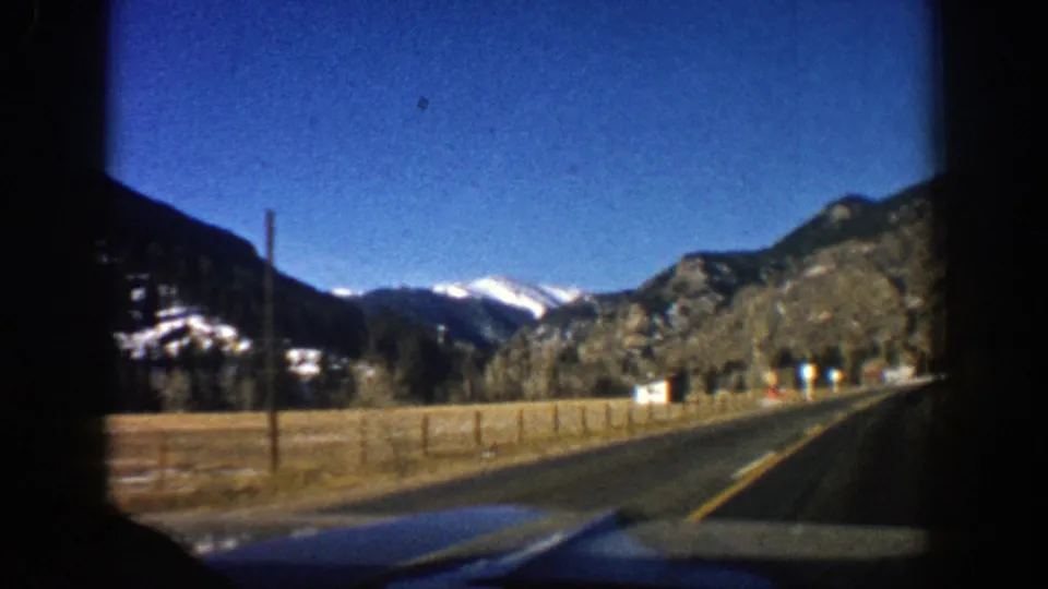 1961 Aspen-area driving view along an open road bordered by fencing, rock slopes, and distant snow-capped peaks.